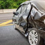 Black sedan heavily damaged on the passenger side after a crash on a highway, with crushed doors and exposed rear taillight assembly.