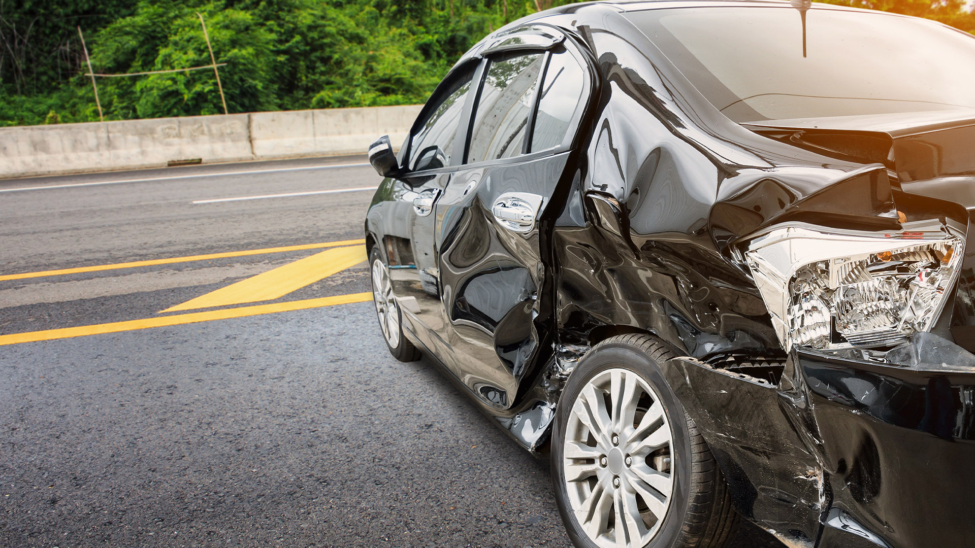 Black sedan heavily damaged on the passenger side after a crash on a highway, with crushed doors and exposed rear taillight assembly.
